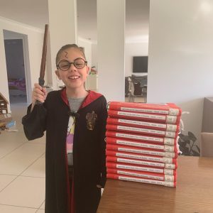 Small girl with a cape and a wand and a lightning scar on her forehead standing next to a braille copy of Harry Potter