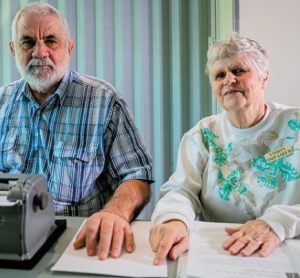 2 people sitting next to each other, one is reading a Braille book and has a perkins machine.
