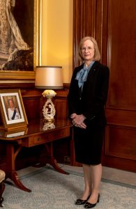 The Governor standing in front of a desk with a lamp and a framed picture of Queen Elizabeth the 2nd