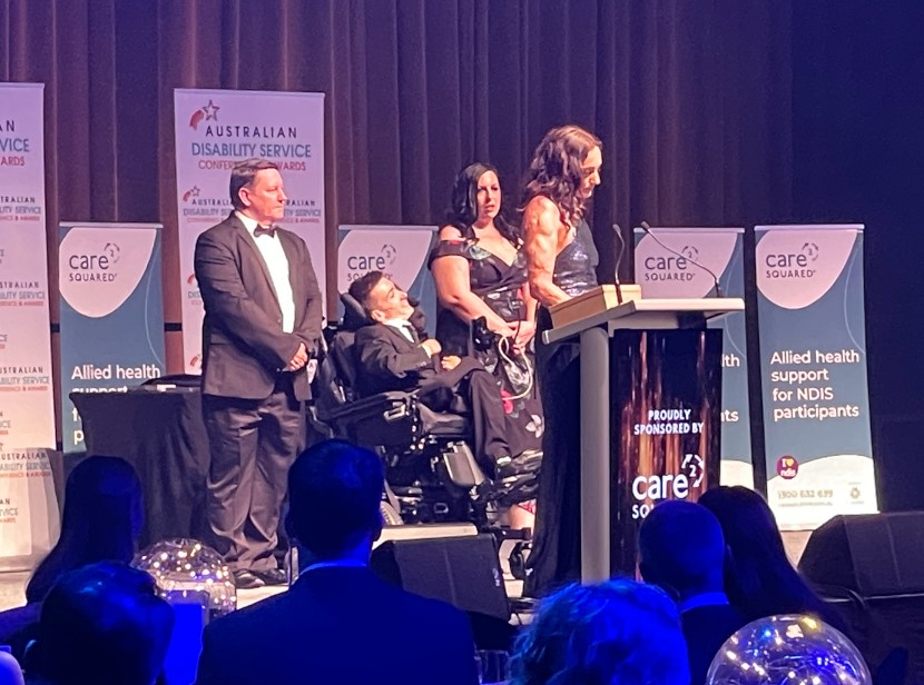 4 people on a stage - one in a wheelchair and one in front of a lectern.
Awards announcement