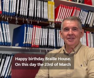 Picture of a man in front of a bookshelf filled with braille books and the text Happy Birthday Braille House.